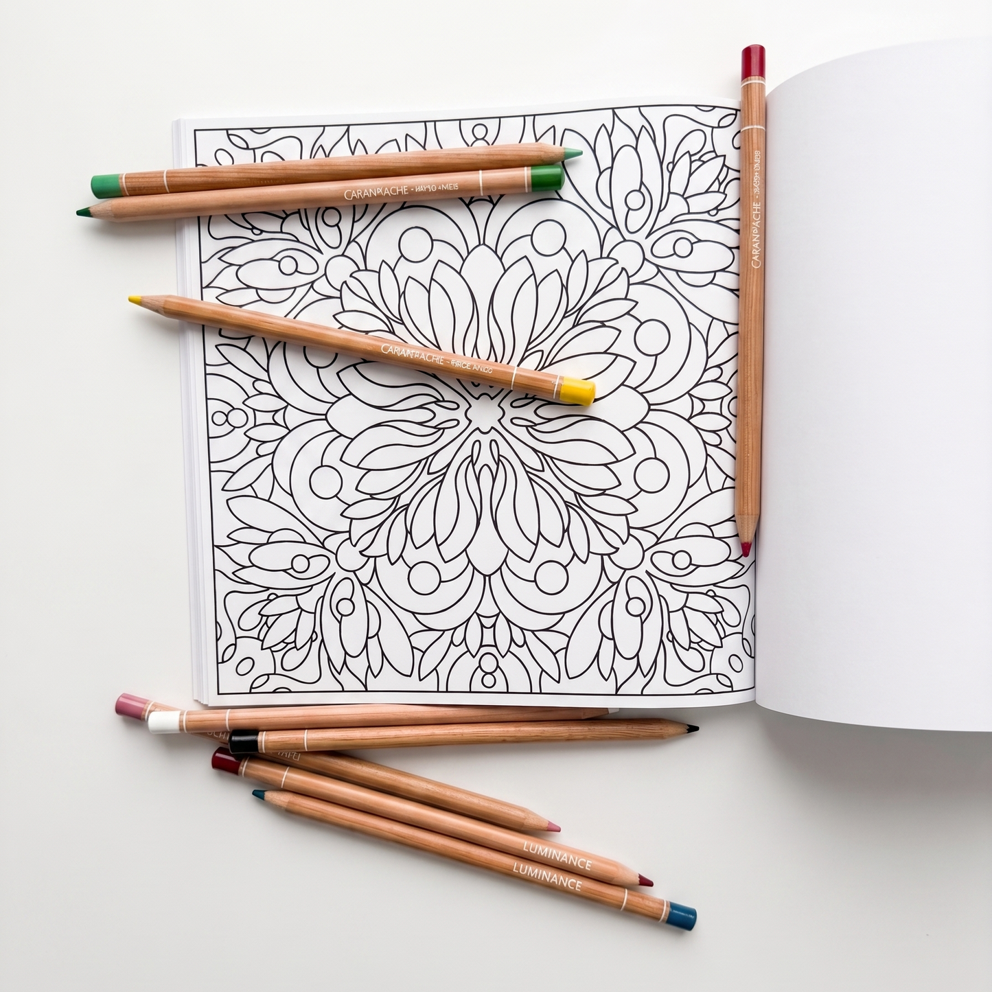 A top-down photograph of an open coloring book with an intricate black and white mandala. Scattered over the page are a set of six Caran d'Ache Luminance colored pencils with a wood finish, with some loose pencils in the foreground and a few partially colored in areas on the page. The background is a plain off-white surface.