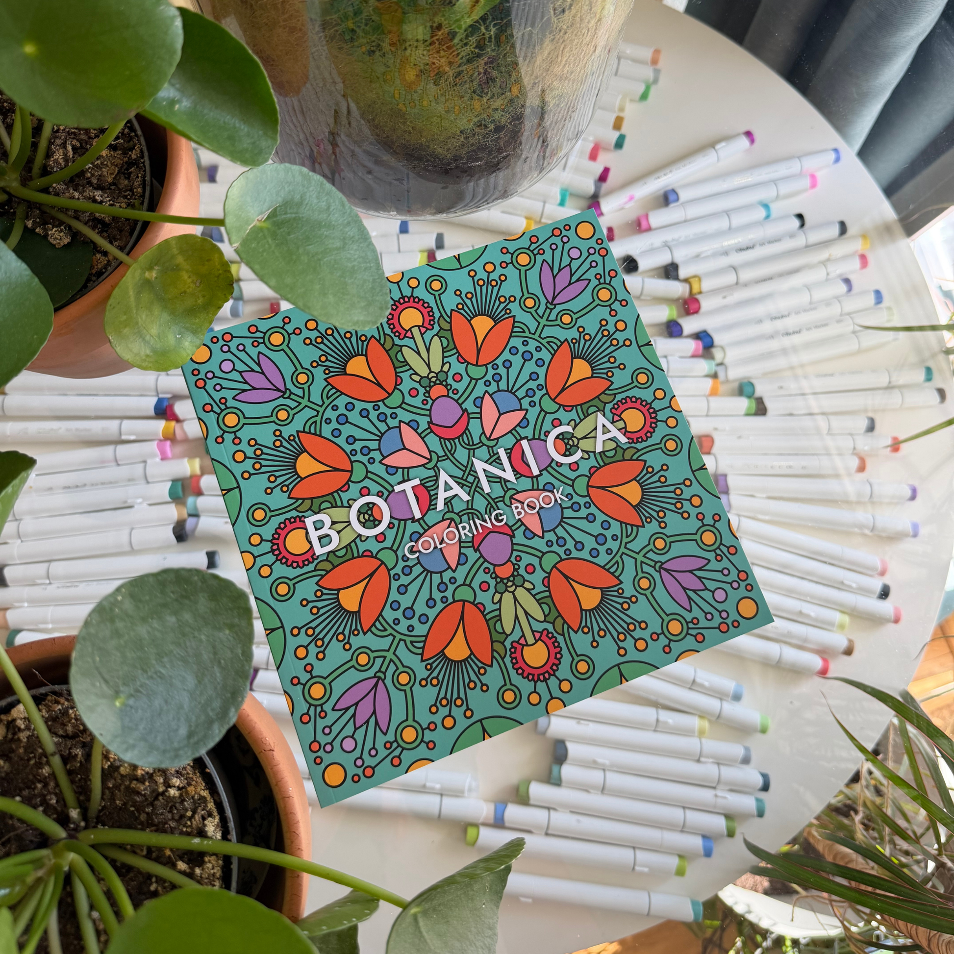 Overhead flat lay of the BOTANICA coloring book cover surrounded by green house plants and colorful art markers.