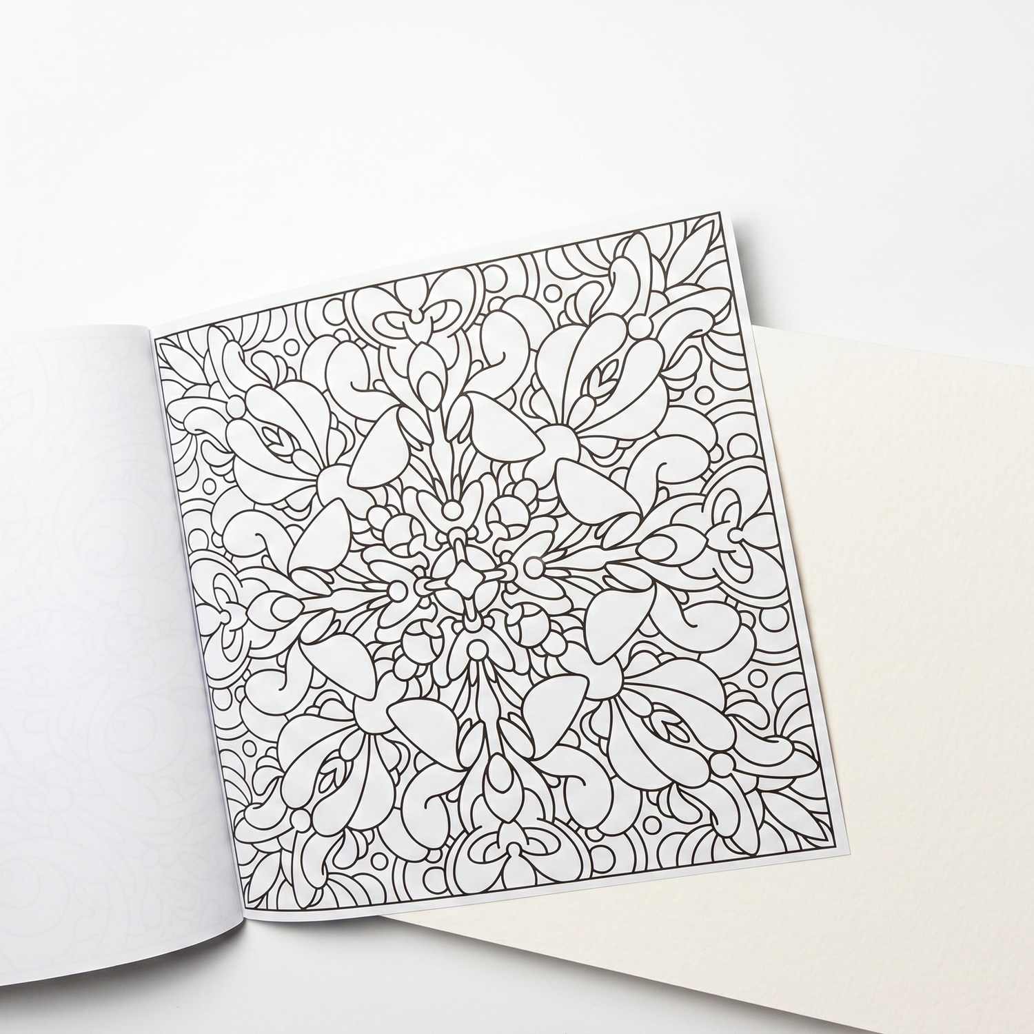 Open coloring book with intricate floral mandala line art rests on a textured cream-colored insert sheet.