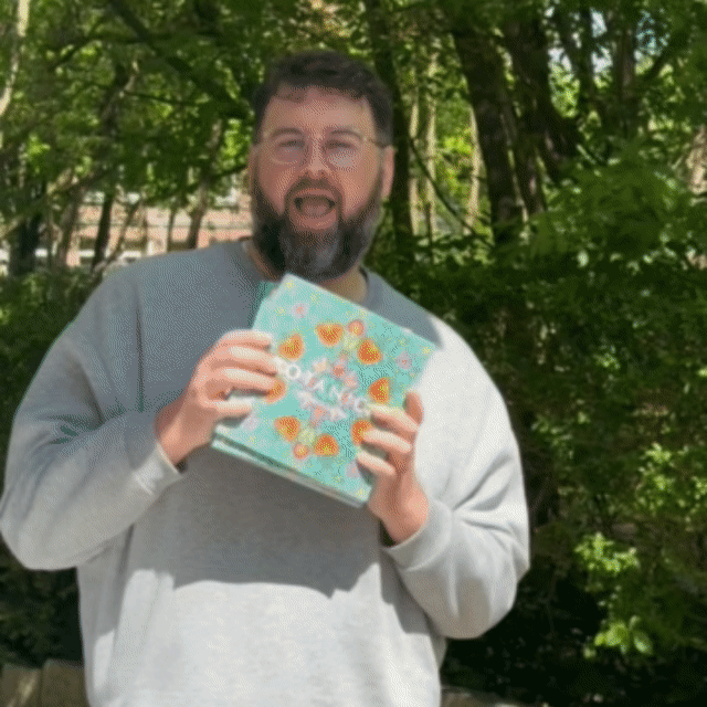 Man jumping up and down from joy, holding BOTANICA coloring books in his hands, outdoors in a park